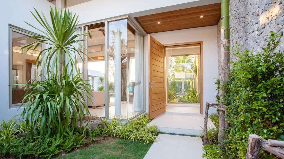 A close-up of the front of a white house with a light-wooden door and floor to ceiling windows, with a palm tree and plants lining the entrance.